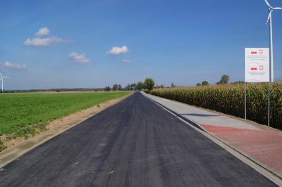 Droga asfaltowa, po lewej stronie pas zieleni, po prawej stronie chodnik i tablica informacyjna